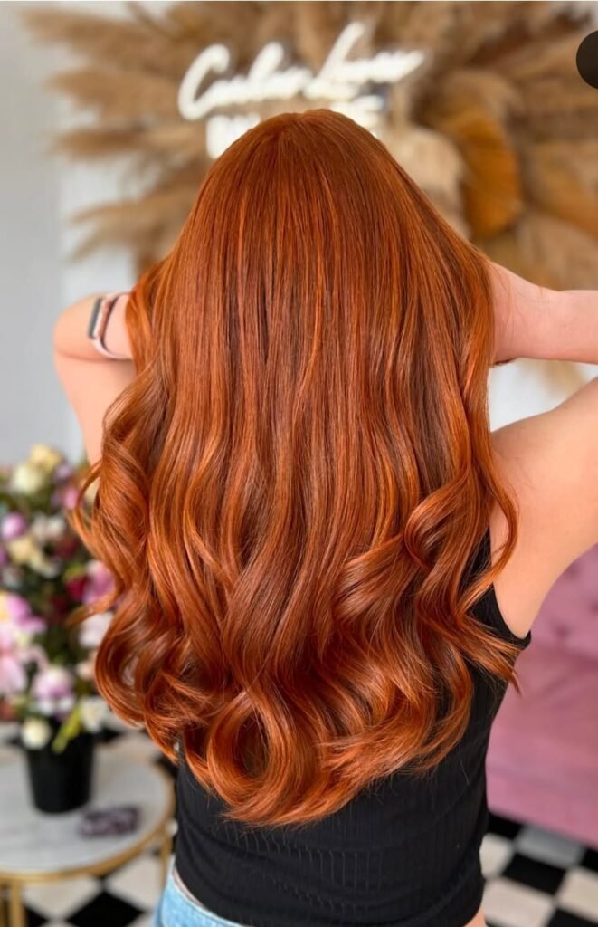 Mujer de espaldas con cabello largo, ondulado y teñido de un tono rojo cobrizo vibrante. El fondo incluye decoración floral y un letrero iluminado.